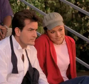 Um homem sério, de camisa branca e gravata desfeita, senta-se ao lado de uma mulher sorridente com boné e casaco vermelho. Cenário exterior.