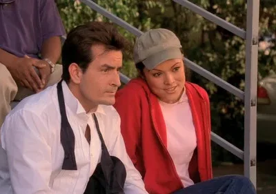 Um homem sério, de camisa branca e gravata desfeita, senta-se ao lado de uma mulher sorridente com boné e casaco vermelho. Cenário exterior.