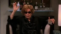 Mulher de óculos escuros e robe preto, fumando cigarro e segurando drink. Senta em poltrona, bar com garrafas ao fundo.