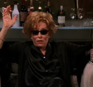 Mulher de óculos escuros e robe preto, fumando cigarro e segurando drink. Senta em poltrona, bar com garrafas ao fundo.