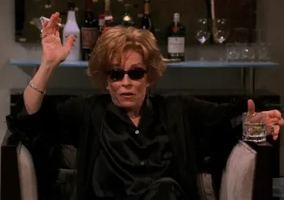 Mulher de óculos escuros e robe preto, fumando cigarro e segurando drink. Senta em poltrona, bar com garrafas ao fundo.