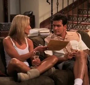 Mulher loira sorri mostrando um papel a um homem, ambos relaxando no sofá com vinho. Cena doméstica casual.