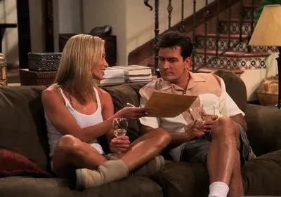 Mulher loira sorri mostrando um papel a um homem, ambos relaxando no sofá com vinho. Cena doméstica casual.