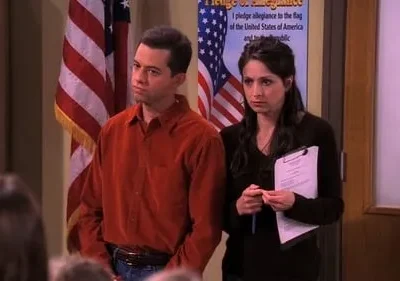 Homem e mulher sérios em sala com bandeira americana. Ele veste laranja, ela, preto com prancheta.