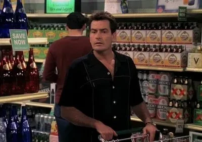 Homem de camisa preta empurra carrinho de compras em corredor de supermercado lotado de bebidas, com expressão surpresa.