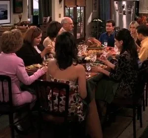 Pessoas reunidas em um jantar festivo, com mesa farta e peru. Cenário de lar aconchegante com cozinha e lareira visíveis.