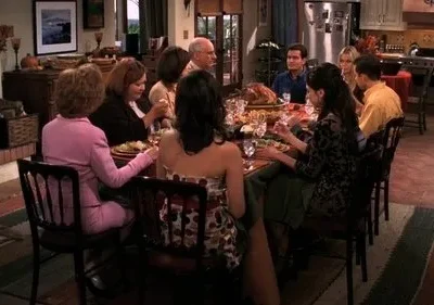 Pessoas reunidas em um jantar festivo, com mesa farta e peru. Cenário de lar aconchegante com cozinha e lareira visíveis.