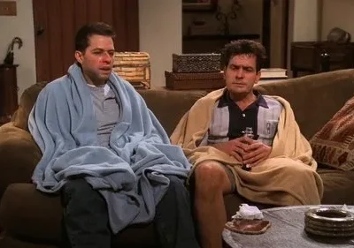 Dois homens parecem doentes ou exaustos, sentados num sofá. Um está envolto em cobertor azul, o outro em bege, segurando uma garrafa.
