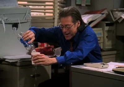 Homem de óculos derrama água de garrafa em copo que vaza, fazendo bagunça em uma mesa de escritório desorganizada.