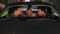 Três pessoas num carro: dois homens nos bancos da frente conversam intensamente, enquanto uma mulher loira os observa do banco de trás.