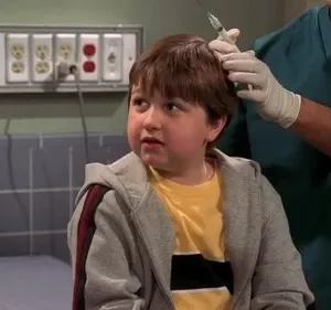 Um menino com expressão apreensiva recebe uma injeção na cabeça de um profissional de saúde com luvas, em consultório médico.