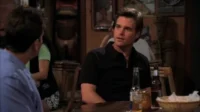 Dois homens em um bar com pouca luz. Um de camisa preta olha para o outro, de costas, em um ambiente descontraído.