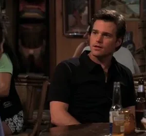 Dois homens em um bar com pouca luz. Um de camisa preta olha para o outro, de costas, em um ambiente descontraído.