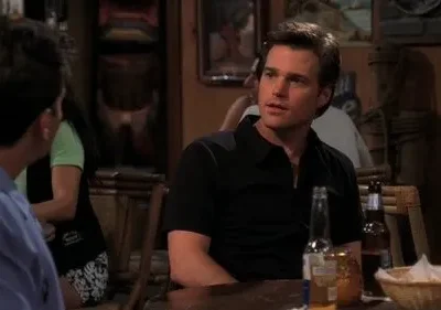 Dois homens em um bar com pouca luz. Um de camisa preta olha para o outro, de costas, em um ambiente descontraído.