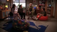 Quatro pessoas conversam numa sala aconchegante. Dois homens e duas mulheres sentam em cadeiras e pufes coloridos, ambiente casual.