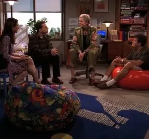 Quatro pessoas conversam numa sala aconchegante. Dois homens e duas mulheres sentam em cadeiras e pufes coloridos, ambiente casual.