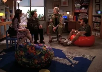 Quatro pessoas conversam numa sala aconchegante. Dois homens e duas mulheres sentam em cadeiras e pufes coloridos, ambiente casual.