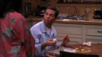 Homem de camisa azul, com ovo na mão, olha perplexo para pessoa. Cozinha com ovos e cascas quebradas na mesa. Clima cômico.