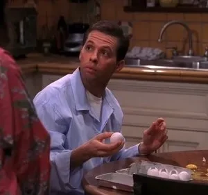Homem de camisa azul, com ovo na mão, olha perplexo para pessoa. Cozinha com ovos e cascas quebradas na mesa. Clima cômico.