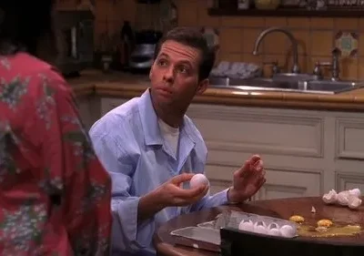 Homem de camisa azul, com ovo na mão, olha perplexo para pessoa. Cozinha com ovos e cascas quebradas na mesa. Clima cômico.