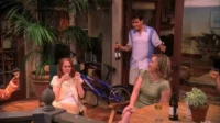 Um homem entra com garrafas em um pátio. Duas mulheres sentadas relaxam com bebidas, uma sorri. Clima descontraído e social.