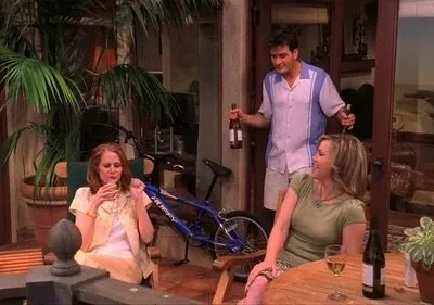 Um homem entra com garrafas em um pátio. Duas mulheres sentadas relaxam com bebidas, uma sorri. Clima descontraído e social.