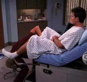 Homem em bata hospitalar deitado em maca de exame, com a perna em estribo, enquanto um profissional se ajoelha aos pés. Ambiente médico.