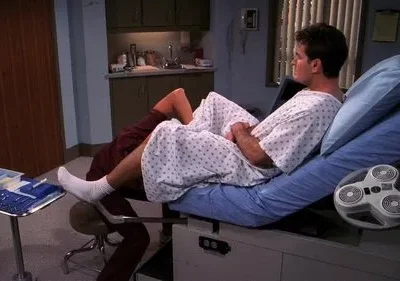 Homem em bata hospitalar deitado em maca de exame, com a perna em estribo, enquanto um profissional se ajoelha aos pés. Ambiente médico.