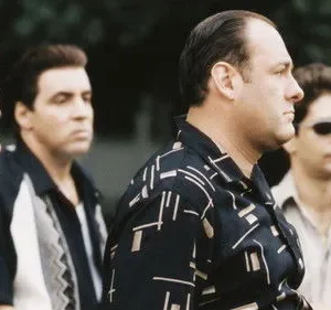 Quatro homens sérios, de meia-idade, com camisas casuais olham para a direita. Um usa óculos escuros. Fundo escuro.