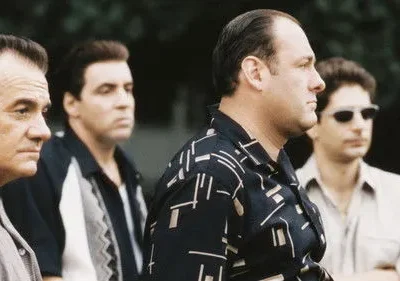 Quatro homens sérios, de meia-idade, com camisas casuais olham para a direita. Um usa óculos escuros. Fundo escuro.