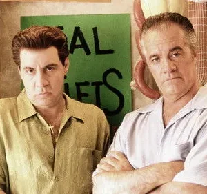 Dois homens de meia-idade, com expressões sérias, posam. Um com camisa verde, outro azul, braços cruzados. Ao fundo, um açougue com salsichas penduradas.