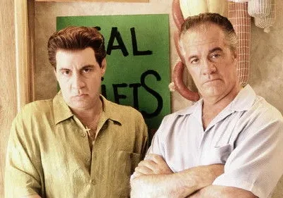 Dois homens de meia-idade, com expressões sérias, posam. Um com camisa verde, outro azul, braços cruzados. Ao fundo, um açougue com salsichas penduradas.