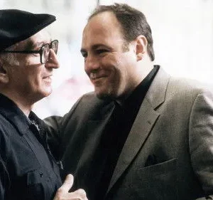 Dois homens sorrindo. O mais velho, de boina, e o mais jovem, de terno cinza com a mão no ombro do outro. Clima amigável.