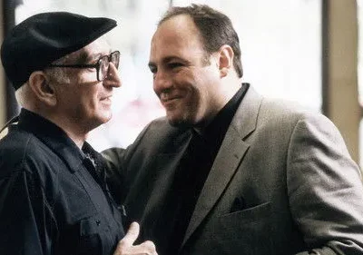 Dois homens sorrindo. O mais velho, de boina, e o mais jovem, de terno cinza com a mão no ombro do outro. Clima amigável.