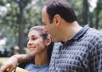 Um homem sorrindo com o braço em volta de uma jovem sorridente. Cenário de parque verde ao fundo. Uma cena feliz e afetuosa.