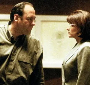 Um homem e uma mulher se encaram intensamente. Ele veste camisa escura, ela blazer claro e óculos. Ambiente sério.