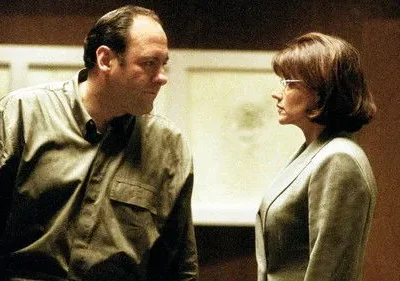 Um homem e uma mulher se encaram intensamente. Ele veste camisa escura, ela blazer claro e óculos. Ambiente sério.