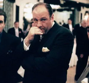 Três homens de terno escuro em um evento formal. O central olha pensativo, com a mão no queixo. Os outros estão sérios. Atmosfera sombria e tensa.