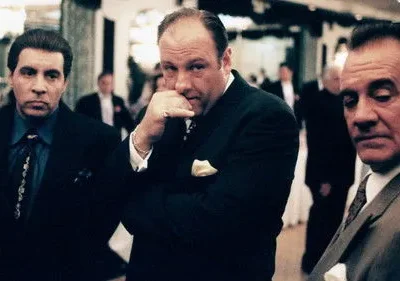 Três homens de terno escuro em um evento formal. O central olha pensativo, com a mão no queixo. Os outros estão sérios. Atmosfera sombria e tensa.