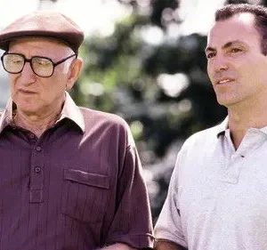 Dois homens em um parque. Um idoso de óculos e boina olha para baixo, o mais jovem ao lado sorri sutilmente.