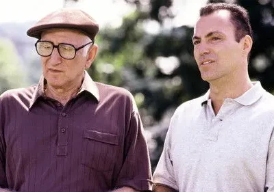 Dois homens em um parque. Um idoso de óculos e boina olha para baixo, o mais jovem ao lado sorri sutilmente.