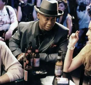 Homem de chapéu sorri para mulher loira fumando, enquanto outro homem observa. Cena de bar noturno com garrafas de cerveja.
