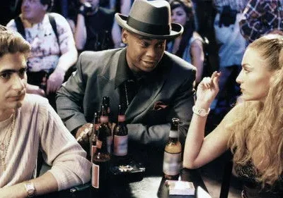 Homem de chapéu sorri para mulher loira fumando, enquanto outro homem observa. Cena de bar noturno com garrafas de cerveja.