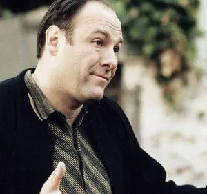 Homem de meia-idade, robusto, com jaqueta escura e camisa listrada, gesticula levemente. Fundo exterior desfocado. Tom sério.