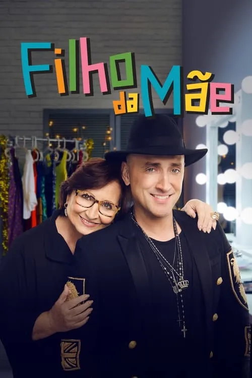 Mulher sorridente de óculos abraça homem de chapéu e blazer preto. Ambos sorriem em um camarim festivo com roupas e luzes.