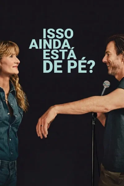 Mulher loira sorri para homem barbudo sorridente com microfone. Fundo escuro. Clima leve e de interação.