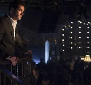 Homem de terno escuro e camisa branca, apoiado em parapeito, observa um clube noturno iluminado por pequenas luzes.