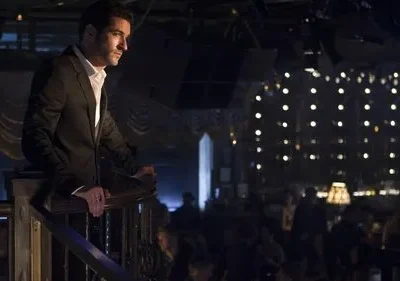 Homem de terno escuro e camisa branca, apoiado em parapeito, observa um clube noturno iluminado por pequenas luzes.