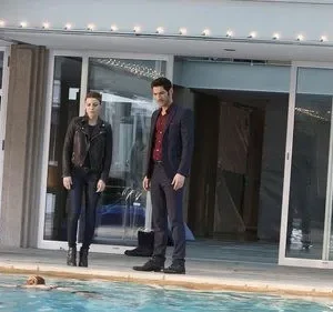 Homem de terno e mulher de jaqueta preta observam uma pessoa flutuando na piscina de uma casa moderna. Clima tenso.