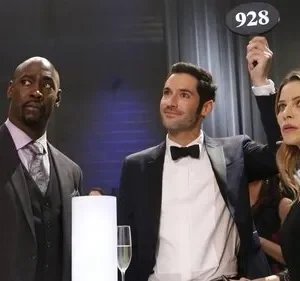 Três pessoas em um evento formal. A mulher segura um número de leilão 928 enquanto os três olham para cima com expressões variadas.